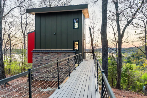 Modern two-story house with a wooden walkway, surrounded by trees and a view of a lake at sunset.