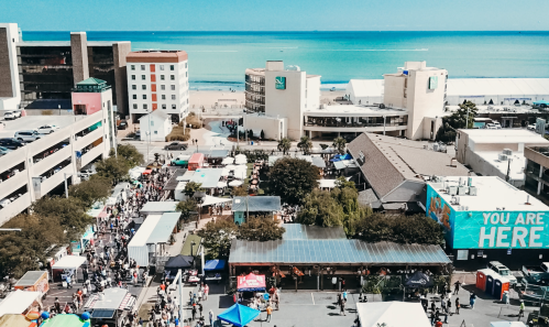 Aerial view of a bustling beachside market with colorful tents, surrounded by buildings and the ocean in the background.