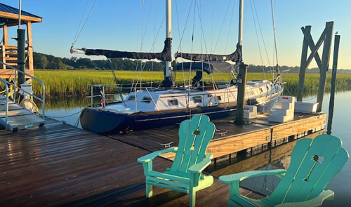 A sailboat docked at a wooden pier with green chairs, surrounded by tall grass and calm water under a clear sky.