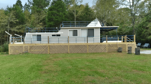 A unique houseboat on a grassy area, featuring a deck and railing, surrounded by trees.