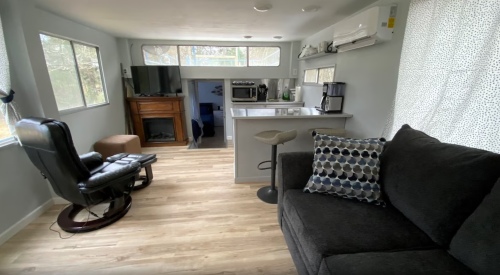 Cozy interior of a small living space with a couch, chair, kitchen area, and large windows for natural light.