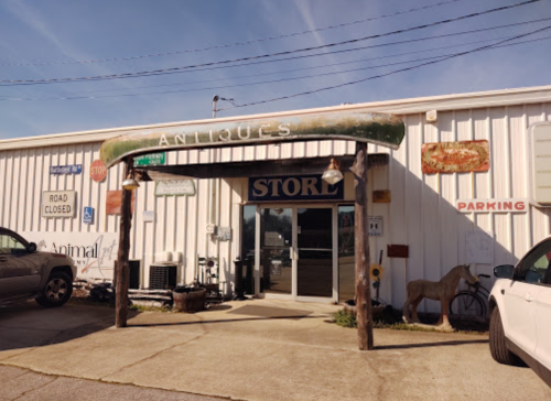 Exterior of an antique store with a sign, parking area, and decorative elements, set against a clear blue sky.