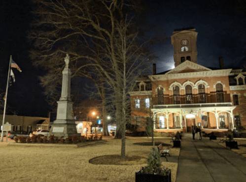 Historic building at night with a clock tower, a monument, and trees, illuminated by streetlights.