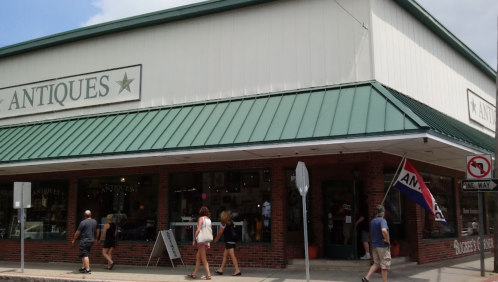 A street view of an antique shop with people walking by and a red, white, and blue flag displayed outside.