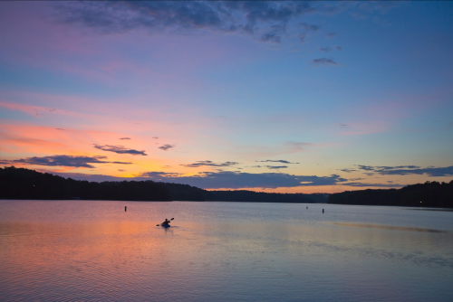 A serene lake at sunset, with a person kayaking under a colorful sky filled with pink and purple hues.