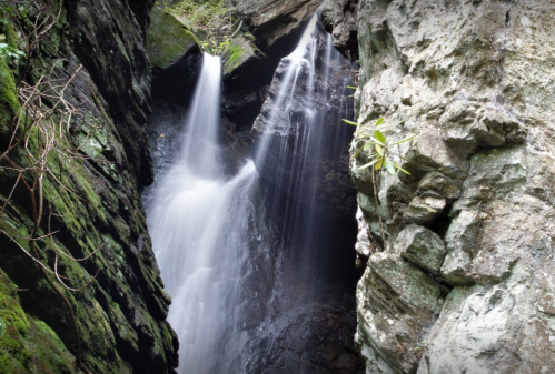 A serene waterfall cascading between moss-covered rocks in a lush, green landscape.