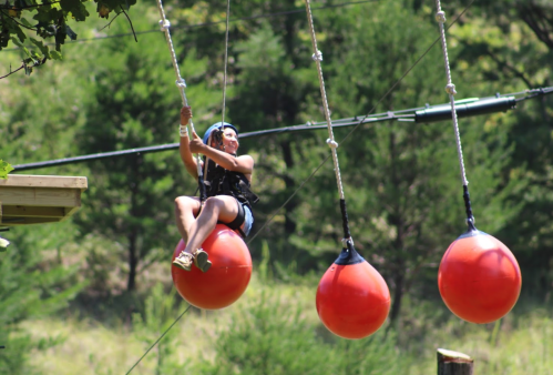 A person swings on a large red ball, enjoying an outdoor adventure activity in a green, wooded area.