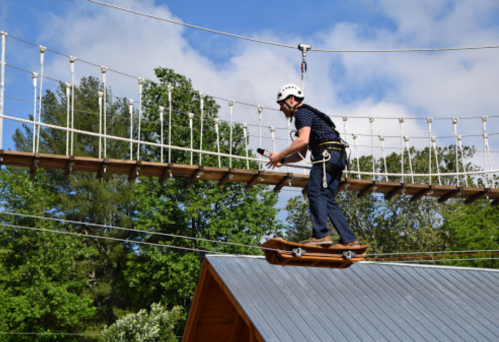 A person in a helmet balances on a skateboard while crossing a rope bridge above a tree-filled landscape.