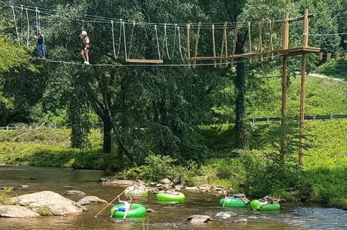 Two people on a rope bridge above a river, while others relax on green inner tubes in the water below.