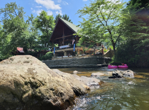 A scenic view of a river with a stone foreground and a wooden cabin surrounded by trees in the background.