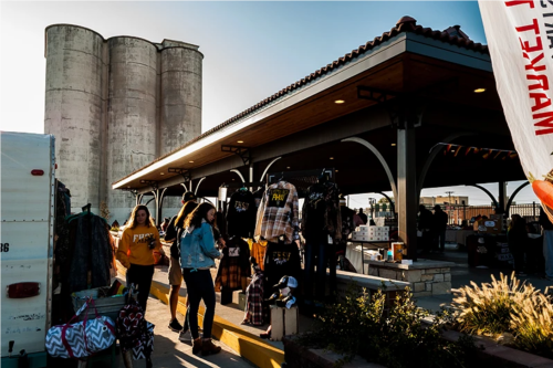 A bustling market scene with people shopping under a covered area near large silos.