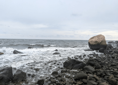 A rocky shoreline with waves crashing against the stones under a cloudy sky. A large boulder is visible in the distance.