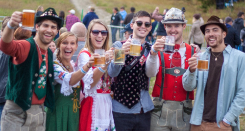 A group of six people in traditional attire cheer with beer mugs at an outdoor festival.