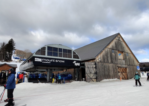 Ski resort scene featuring a ski lift, wooden barn, and skiers on snow under a cloudy sky.