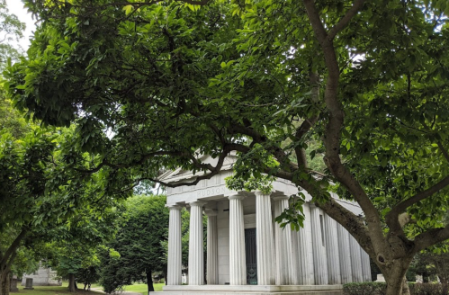 A classical stone structure with columns, partially obscured by lush green trees and foliage.