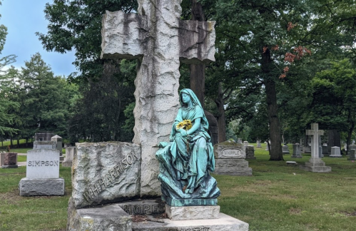 A bronze statue of a seated woman holding flowers beside a large stone cross in a cemetery.