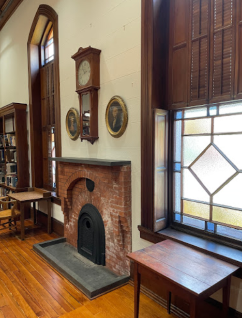 A cozy library interior featuring a brick fireplace, wooden furniture, and decorative stained glass windows.