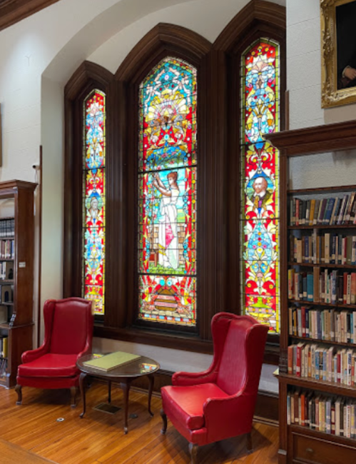 Two red armchairs and a small table in a library, featuring colorful stained glass windows in the background.
