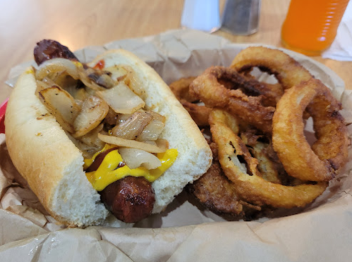 A hot dog topped with grilled onions and mustard, served with a side of crispy onion rings on a paper tray.