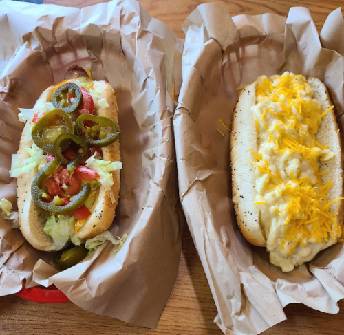 Two hot dogs in paper trays: one topped with jalapeños, lettuce, and tomatoes; the other with cheese sauce and shredded cheese.