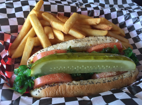 A hot dog topped with lettuce, tomato, and a pickle, served with a side of golden French fries in a checkered basket.