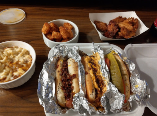 A takeout meal featuring three hot dogs in foil, tater tots, chicken wings, and a bowl of cheesy pasta.