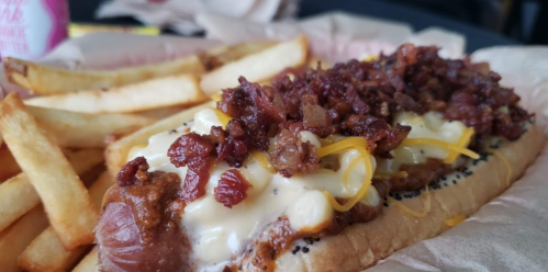 A loaded hot dog topped with cheese, bacon bits, and served with crispy French fries on the side.