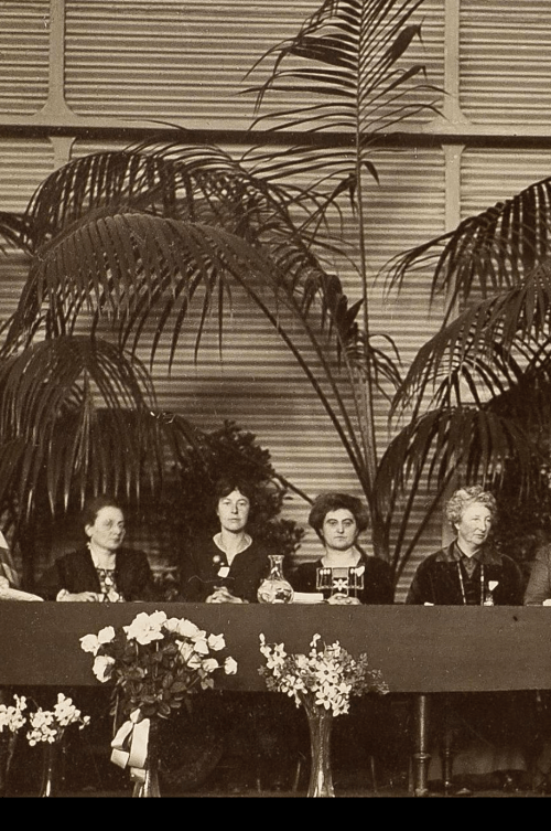 A historical gathering of women seated at a table, with plants and flowers in the background.