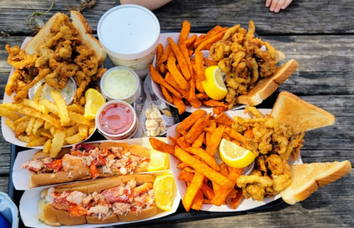 A large platter of seafood, including fried clams, lobster rolls, sweet potato fries, and lemon wedges on a wooden table.