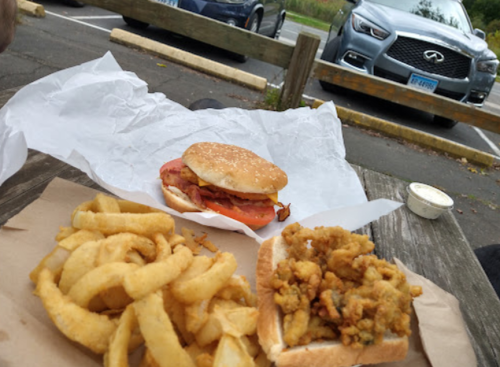 A sandwich with bacon and tomato next to a tray of fries and a serving of fried seafood, with cars in the background.
