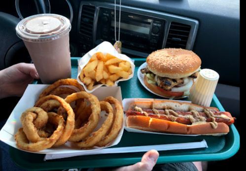 A tray of fast food featuring onion rings, French fries, a burger, and a hot dog with a chocolate milkshake.