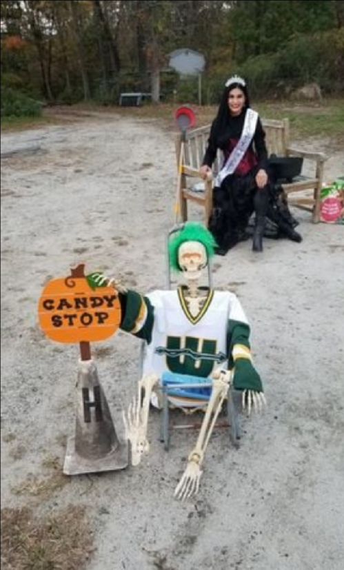 A skeleton in a hockey jersey holds a "CANDY STOP" sign, while a woman in a costume sits nearby on a bench.