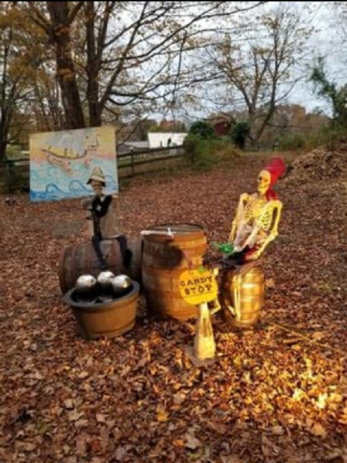 A skeleton and a figure in a hat sit by barrels in a fall setting, with a sign reading "Candy Stop" and autumn leaves around.