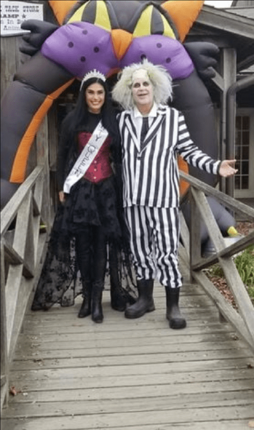 A woman in a black dress and tiara stands next to a man in a striped suit and white wig, in front of a Halloween decoration.