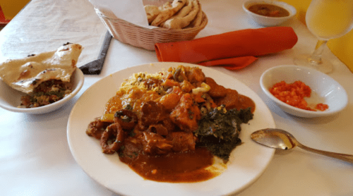 A plate of colorful curry with vegetables and meat, served with naan, salad, and a small bowl of sauce.