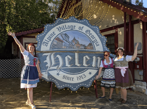 Three women in traditional attire pose beside a large sign that reads "Helen" in the Alpine Village of Georgia.