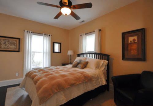 Cozy bedroom with a large bed, ceiling fan, two windows, and a chair, featuring warm tones and decorative artwork.
