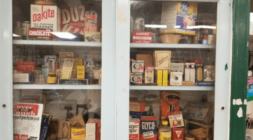 A vintage display cabinet filled with various old cleaning products, food items, and household goods.