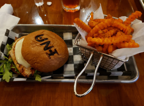 A burger with "UNI" stamped on the bun, served with a side of crinkle-cut sweet potato fries in a small basket.