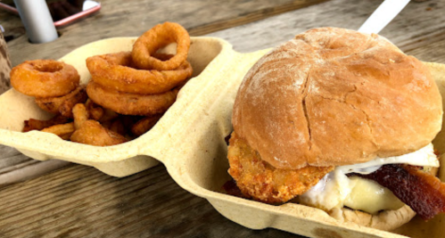 A sandwich with a fried egg, bacon, and a crispy patty, served with a side of onion rings in a takeout container.