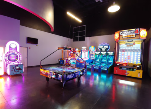 A vibrant arcade featuring various games, including air hockey, skee ball, and a Connect 4 machine, all illuminated with colorful lights.