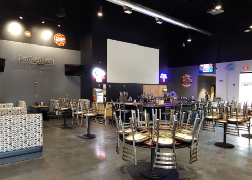 Interior of a restaurant with empty tables and chairs, a bar area, and neon signs on the walls.