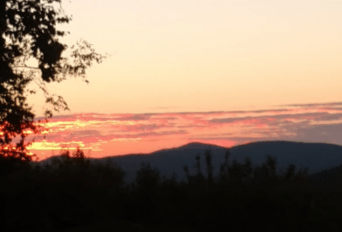 A serene sunset over rolling hills, with vibrant orange and pink hues in the sky and silhouettes of trees in the foreground.