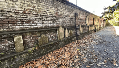 A weathered brick wall lined with old gravestones, surrounded by fallen leaves and a gravel path.