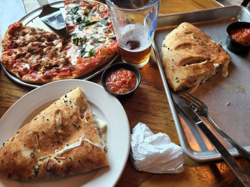 A table with a pizza, a calzone, a beer, and marinara sauce, with a napkin and utensils nearby.