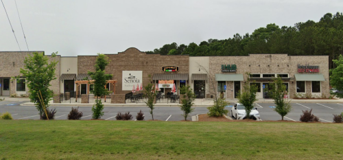A row of storefronts with various businesses, surrounded by green grass and trees under a cloudy sky.
