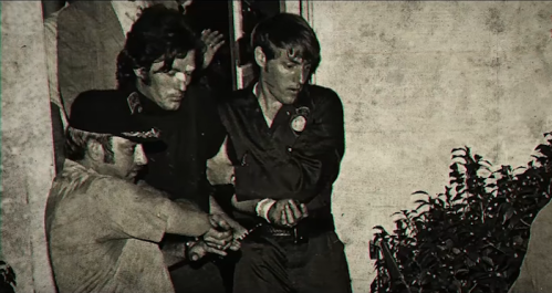 A black-and-white image of two men being escorted by police, one in handcuffs, near a building entrance.