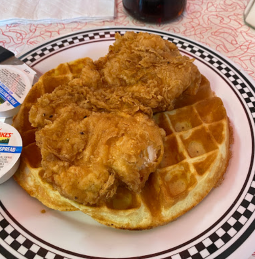 A plate with two pieces of fried chicken on a waffle, accompanied by packets of syrup and spread.