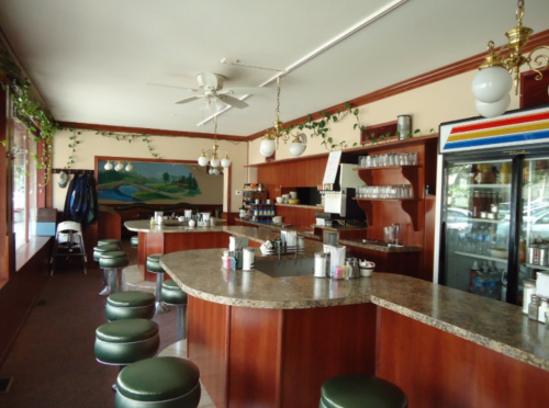 A cozy diner interior featuring a curved bar, green stools, and decorative plants along the walls.