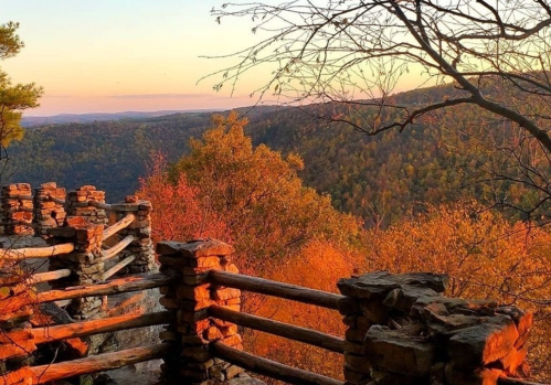 Scenic view of a sunset over a valley, framed by trees and a rustic wooden railing. Warm autumn colors dominate the landscape.